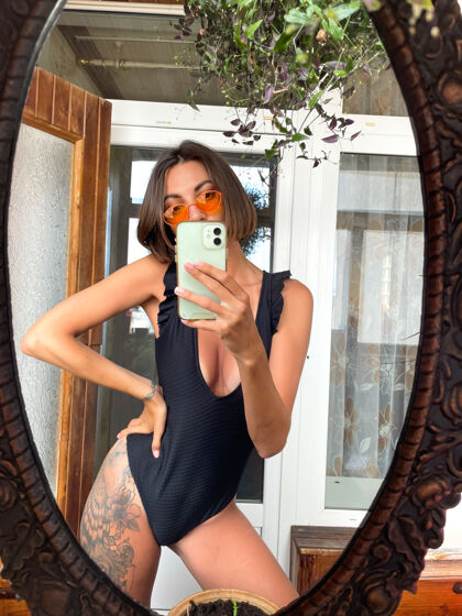 度假家里的美女穿着黑色夏季泳装 在手机上对着镜子自拍 在社交媒体上发布故事和帖子快乐手机时尚