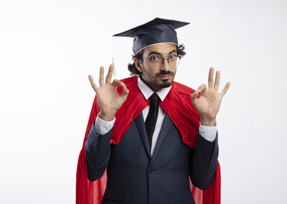 毕业令人印象深刻的年轻白种人超级英雄戴着眼镜 穿着红色斗篷和毕业帽的西装 用两只手做手势表示同意光学复制空间