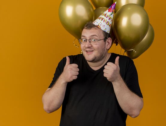 手兴奋的成年斯拉夫人戴着眼镜 戴着生日帽 站在氦气球前 双手竖起大拇指帽子穿男人