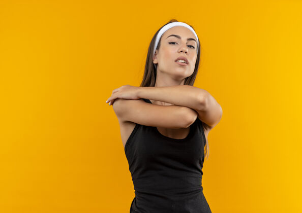年轻自信的年轻漂亮的运动女孩戴着头带和手腕交叉双臂在橙色的墙上头带运动腕带