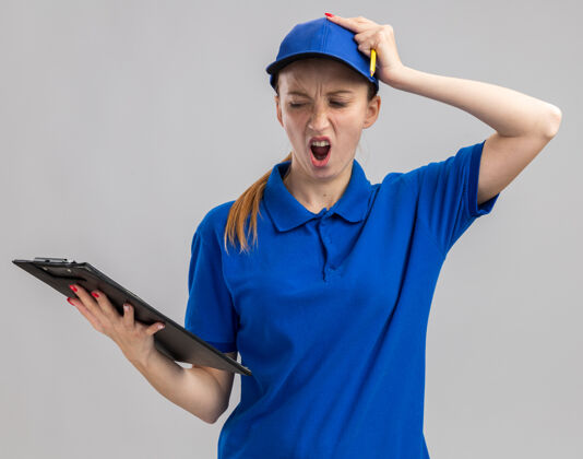 手穿着蓝色制服 戴着帽子的年轻送货女孩拿着剪贴板 困惑地看着它 手放在头上 因为她站在白色的墙上犯了错误帽子剪贴板错误