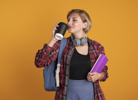 笔记本高兴的年轻斯拉夫女学生戴着耳机背着书包拿着纸杯书和笔记本空间橙色纸