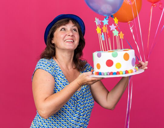 举行快乐的中年妇女戴着派对帽 手里拿着五颜六色的气球 举着生日蛋糕 脸上挂着微笑 站在粉色的墙上庆祝生日派对气球脸微笑