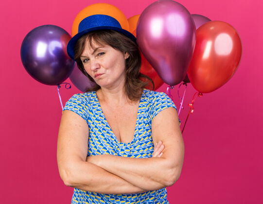 五颜六色中年妇女戴着派对帽 手里拿着五颜六色的气球 不高兴地交叉双臂站在粉色的墙上庆祝生日派对举行中间帽子