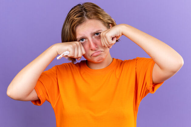 可怕不健康的年轻女子穿着橙色t恤手握纸巾感觉很难受流鼻涕咳嗽感冒哭泣用力揉眼睛站在紫色的墙上年轻女人抱着