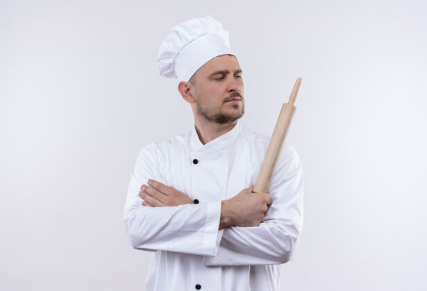 卷自信的年轻帅哥厨师身着厨师制服 站在那里 紧闭的姿势 拿着擀面杖 看着隔离在白墙上的一面别针烹饪姿态