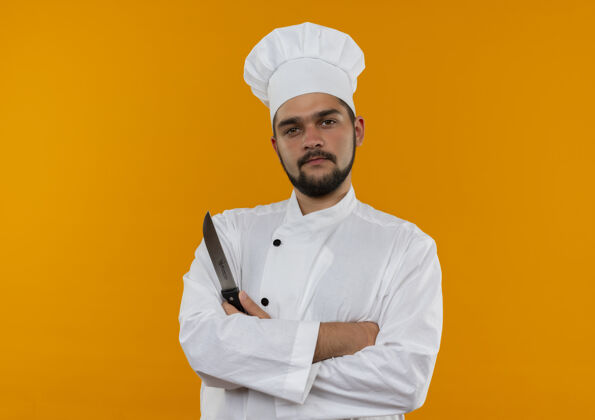姿势自信的年轻男性厨师身着厨师制服 站在封闭的姿势 手持隔离在橙色墙壁上的刀 留有复印空间厨师封闭刀