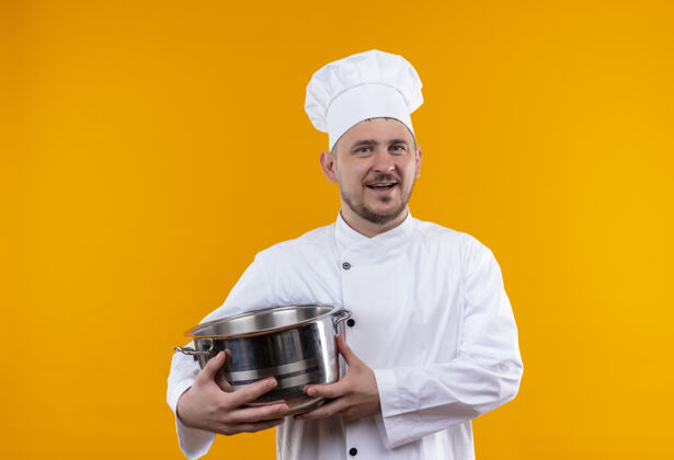 烹饪快乐的年轻帅哥厨师穿着厨师制服 拿着孤立在橙色墙上的锅手持年轻制服
