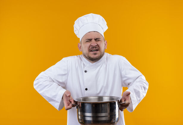 年轻恼怒的年轻帅哥厨师穿着厨师制服拿着锅炉闭着眼睛站在孤立的橙色墙上封闭锅炉持有