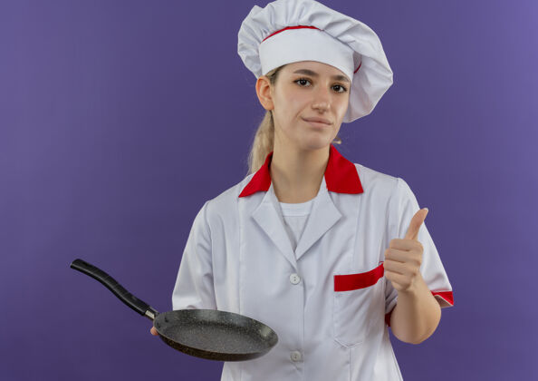 拇指自信的年轻漂亮厨师 身着厨师制服 手拿煎锅 在紫色的墙上孤立地竖起大拇指薯条平底锅拿着