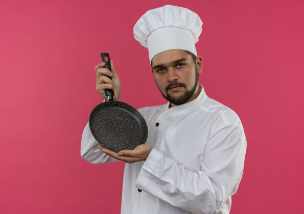 年轻自信的年轻男性厨师身着厨师制服 手拿煎锅 隔离在粉色墙壁上 留有复印空间男性平底锅厨师
