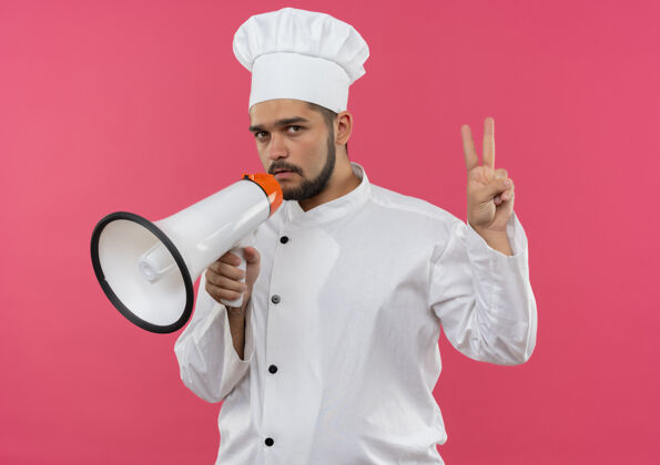 制服自信的年轻男厨师身着厨师制服 手持喇叭 在粉红色的墙上做着和平的手势演讲者年轻男性