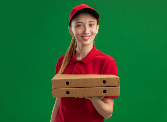 拿着身穿红色制服 头戴鸭舌帽 手持披萨盒的年轻送货女孩站在绿色的墙上 脸上带着幸福的微笑年轻送货制服