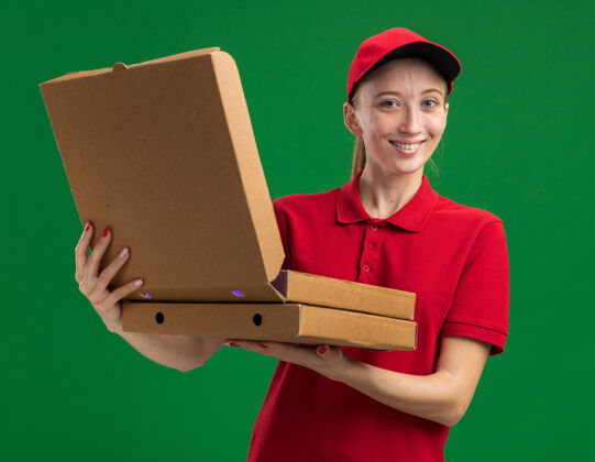 帽子身穿红色制服 头戴鸭舌帽 手持披萨盒打开其中一个披萨盒的年轻送货女孩友好地站在绿色的墙上送货打开披萨
