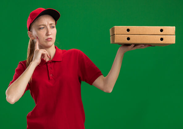 拿着身穿红色制服 头戴鸭舌帽 拿着披萨盒的年轻送货员站在绿色的墙上 困惑地看着披萨盒女孩送货站着