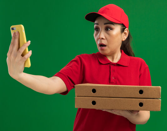 站着年轻的送货员身穿红色制服 头戴鸭舌帽 手里拿着披萨盒 站在绿色的墙上 看着智能手机的屏幕 心神不安制服手机担心