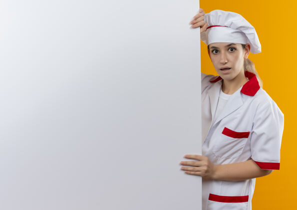 墙年轻漂亮的厨师穿着厨师制服站在身后 拿着白色的墙 隔离在橙色的墙上 留有复印空间印象拿着制服