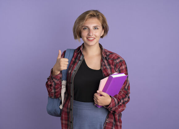 背包微笑着的年轻的斯拉夫学生女孩背着背包 竖起大拇指 拿着书和笔记本紫色笔记本微笑