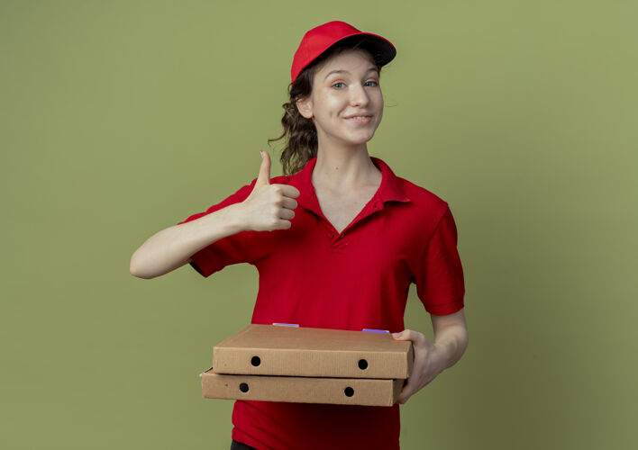 制服高兴的年轻漂亮的送货女孩在红色制服和帽子举行比萨饼包 并显示拇指了送货橄榄女孩