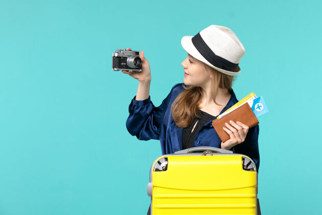 年轻女子前视图：手持相机和机票的年轻女子乘坐蓝色背景的女子海上旅行飞机机票旅行帽子