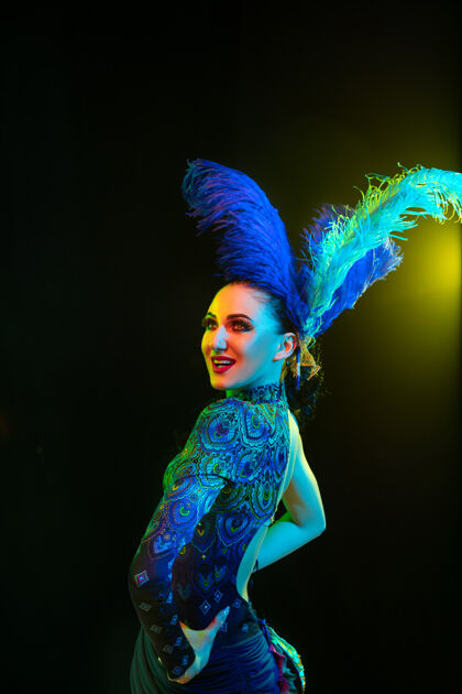剧院嘉年华里美丽的年轻女子 霓虹灯下黑色墙壁上羽毛的时尚化装服装面具脸女人