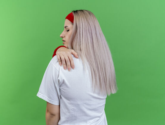 绿色疼痛的年轻白人运动女孩戴着背带 戴着头带和腕带 背对着摄像机 手放在肩膀上疼痛佩戴背部