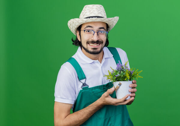 绿色年轻的留着胡须的园丁 穿着连体衣 戴着帽子 手里拿着盆栽植物 站在绿色的背景上 面带微笑 满脸幸福微笑锅穿