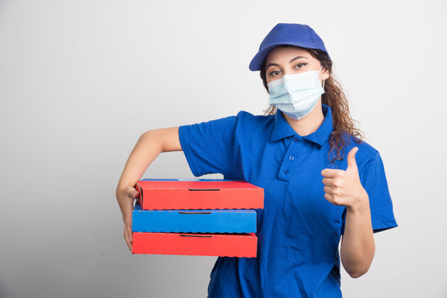 比萨饼送比萨饼的女孩拿着三个盒子 戴着医用口罩 大拇指朝上竖着白色的美味盒子工人
