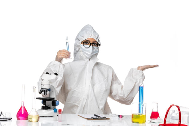 专业正面图身穿特殊防护服的年轻女化学家拿着放在白桌子上的蓝色溶液的烧瓶实验室病毒化学特殊人面具