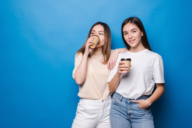 拥抱两个年轻的女人拿着咖啡杯孤零零地走在蓝色的墙上有趣人杯子