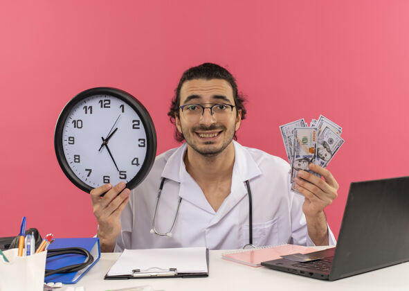 桌子面带微笑的年轻男医生 戴着医用眼镜 穿着医用长袍 听诊器坐在办公桌旁现金空间时钟