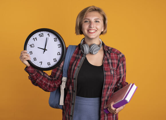 学生微笑的年轻斯拉夫女学生戴着耳机 背着背包 手里拿着钟表本和笔记本橙色背包笔记本