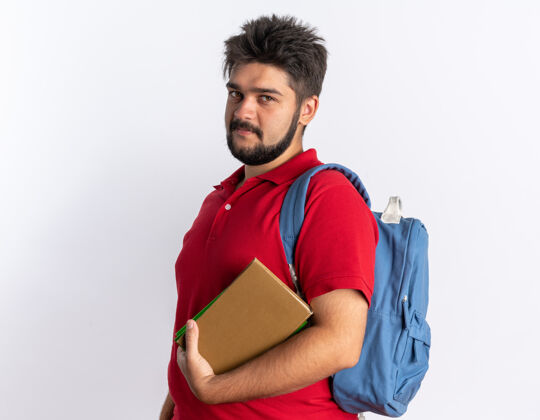 胡子年轻的留着胡子的学生 穿着红色马球衫 背着背包 手里拿着笔记本 站在那里微笑 快乐 积极正面学生站着