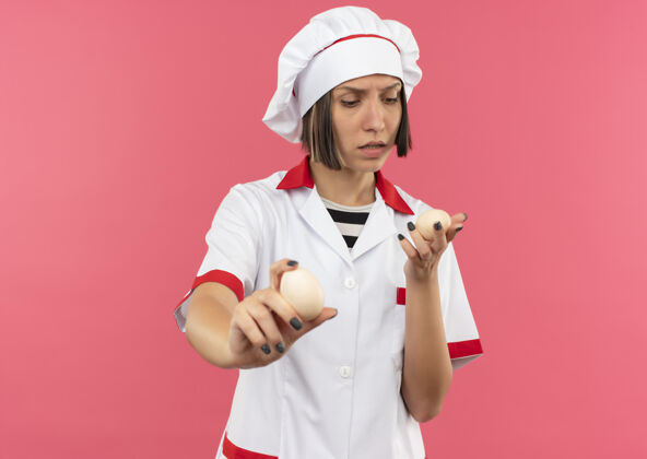 烹饪体贴的年轻女厨师穿着厨师制服 手拿着粉红色墙上孤立的鸡蛋 望着 伸着脸人年轻