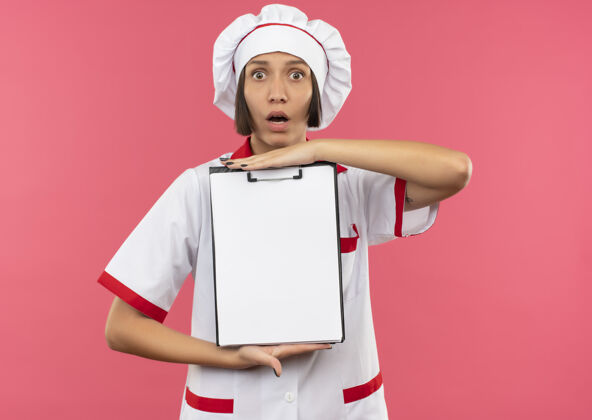市民惊讶的年轻女厨师穿着厨师制服拿着剪贴板看着前面孤立的粉色墙壁制服人人