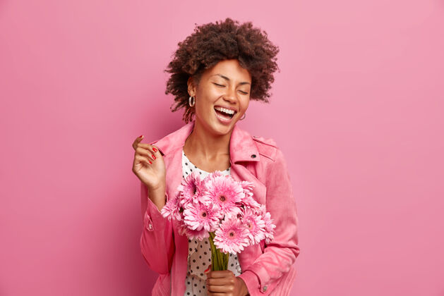 快乐快乐无忧无虑的女人的肖像手持一束非洲菊花表达积极的情绪闭上眼睛穿着时尚的夹克隔离在粉红色的墙上头发乐趣年轻