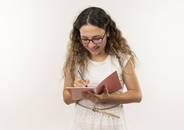 表情带着微笑的年轻漂亮的女学生戴着眼镜背着书包在白色的墙上用钢笔写着笔记人包穿