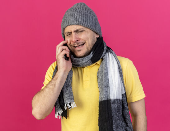 疾病悲伤的年轻金发病斯拉夫男子戴着冬季帽子和围巾在电话上交谈孤立的粉红色围巾男人年轻