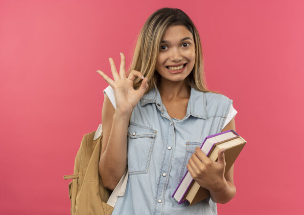脸微笑着的年轻漂亮的学生女孩 背着书包 拿着书 在粉红色的墙上贴着“做得很好”的牌子人Ok微笑