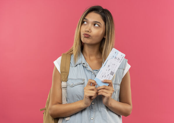 人体贴的年轻漂亮的女学生 背着书包 拿着机票 看着粉红色墙上孤立的一面飞机票人