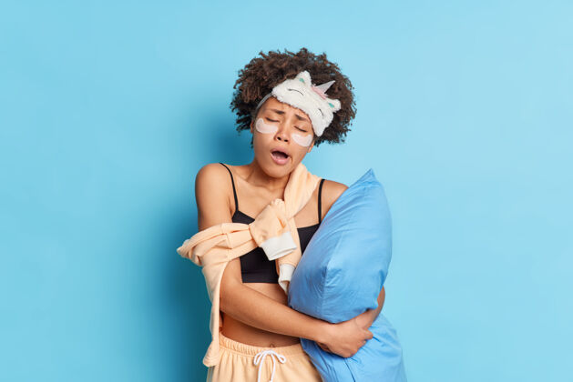 年轻睡意朦胧 卷发的美国黑人妇女早起后打哈欠的画像穿着睡衣抱着枕头 睡眠面罩歪着头 眼睛下面贴着胶原蛋白垫 隔着蓝色的墙可爱休息舒适