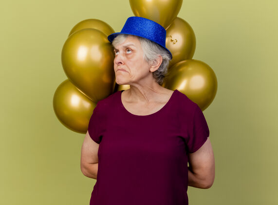 老人困惑的老妇人戴着派对帽 身后拿着氦气球 仰望橄榄绿橄榄女人聚会