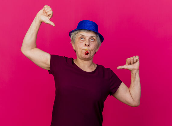 党戴着派对礼帽的自信的老妇人用两只手指着自己吹着粉红色的口哨女人帽子吹