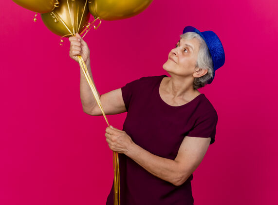 气球高兴的老妇人戴着聚会的帽子站在那里 看着粉红色的氦气球粉色派对女士