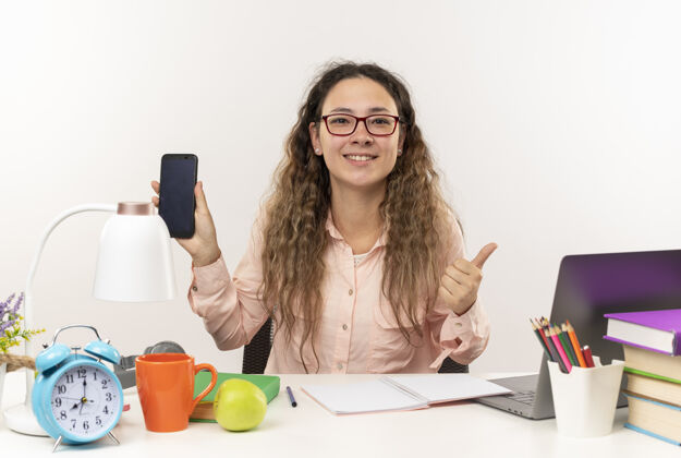 人微笑着的年轻漂亮的女学生戴着眼镜坐在课桌旁 拿着学习用具做作业 把手机和大拇指孤立地竖立在白色的墙上手机白色女生