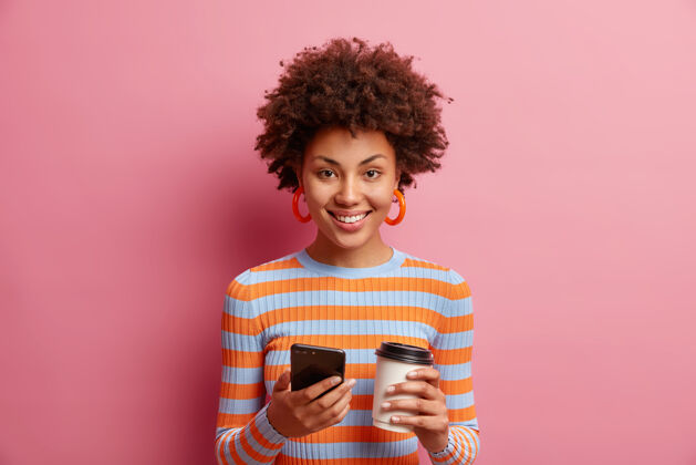 青少年迷人满足的年轻黑人女性使用智能手机进行在线交流有咖啡休息拿着纸杯享受美好的一天穿着条纹套头衫隔着粉色的墙壁跳伞表情快乐