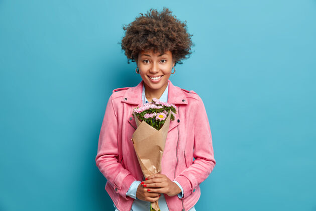室内美丽美丽的非洲裔美国女人 卷发 手持花束 准备祝贺好友 节日快乐 穿着粉色夹克 隔离在蓝色的工作室墙上礼物成人花
