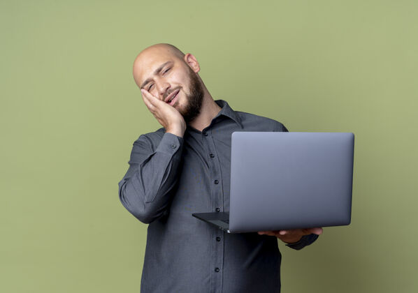 电话疲惫的年轻秃头呼叫中心男子拿着笔记本电脑把手放在脸上隔离在橄榄绿的墙上人橄榄脸