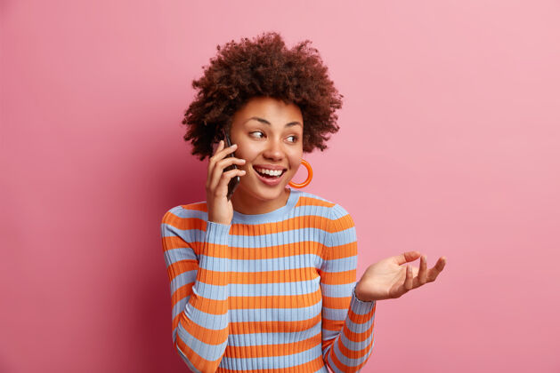 毛衣快乐美丽的非裔美国女人在电话里交谈愉快的交谈看着远方可爱的微笑举起手穿着休闲条纹套头衫隔着粉色的墙壁有有趣的谈话人人表情