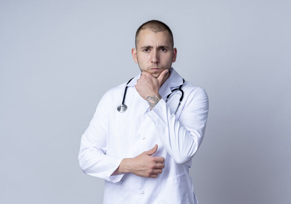 男性体贴的年轻男医生 穿着医用长袍 脖子上戴着听诊器 手放在隔离在白墙上的下巴上手医学人